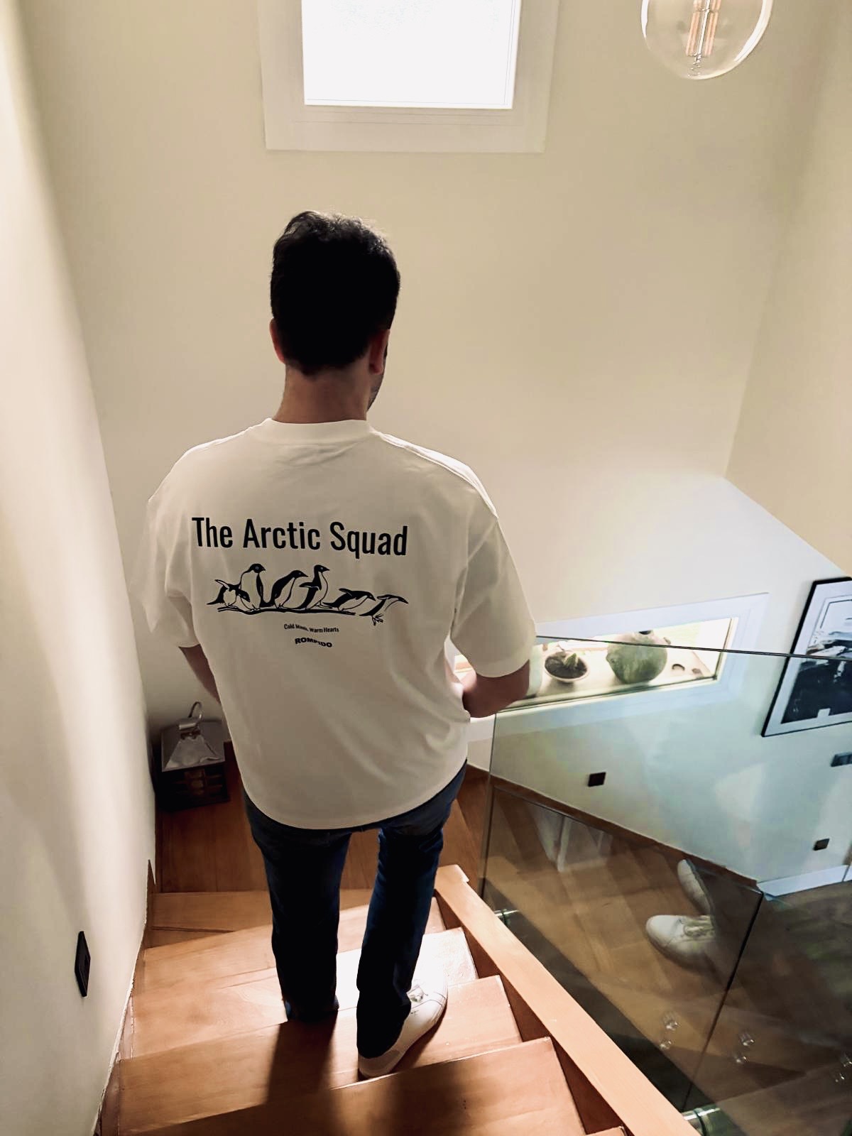 Chico joven moreno de espaldas bajando las escaleras con una camiseta blanca que tiene un dibujo de unos pingüinos con la frase The Arctic Squad